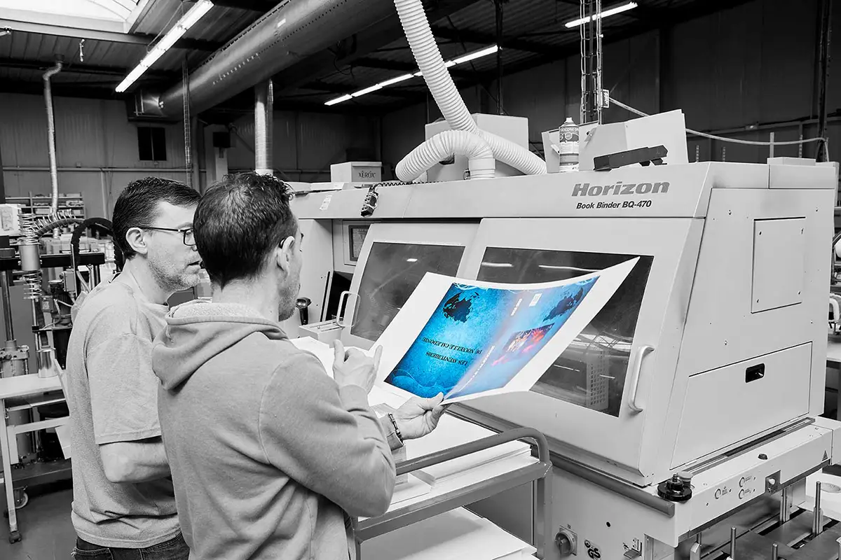 1 homme tenant une feuille de papier bleu discutant avec un collègue devant la relieuse